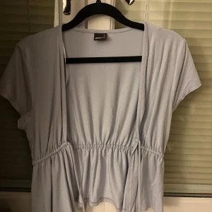 Ljusblå omlottopp från Gina Tricot - Säljer en ljusblå omlottopp från Gina Tricot med korta ärmar och knyt i midjan. Toppen har en avslappnad passform och är tillverkad i mjuk bomull, perfekt för en chill och stilren look.