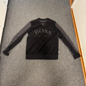 Svart tröja från Hugo Boss - Snygg svart tröja från Hugo Boss med stora BOSS-trycket framtill. Ärmarna är mörkgrå och ger en cool kontrast till resten av tröjan. Mjuk bomullskänsla och klassisk passform med rund hals. Perfekt för dig som gillar stilrena märkeskläder. M passar även S 