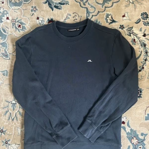 Svart sweatshirt från J.Lindeberg - En stilren svart sweatshirt från J.Lindeberg med rund hals och diskret logga på bröstet. Tröjan har långa ärmar och ribbade muddar vid ärmslut och nederkant. Perfekt för en clean och enkel look.