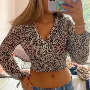 Leopardmönstrad croppad blus från Zara - Säljer en croppad blus från Zara med leopardmönster i svart och beige. Blusen har långa ärmar, v-ringning med volangdetalj och smockad midja som ger en snygg passform. Perfekt för dig som vill sticka ut med ett trendigt djurmönster.