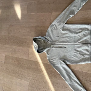 Grå hoodie från Polo Ralph Lauren - Snygg ljusgrå hoodie från Polo Ralph Lauren med dragkedja och huva. Klassisk broderad logga på bröstet, två fickor framtill och justerbar snörning i huvan. Perfekt för en chill och stilren look. Storlek S.