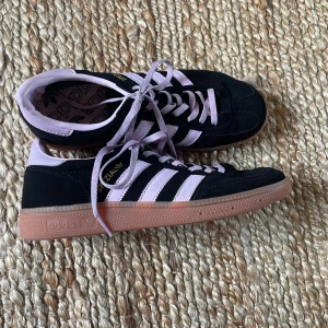 Adidas Spezial sneakers  - Säljer ett par Adidas Spezial sneakers i svart mocka med lila detaljer och klassiska tre ränder i lila/ljusrosa.  Skorna har snörning, gummisula i ljusbrun ton och rund tå. Perfekta för dig som gillar retrostil och vill sticka ut med en unik färgkombo. Storlek 38. Använda ett fåtal gånger därav lite missfärgat på snörena. Skriv för mer bilder eller övriga frågor. 