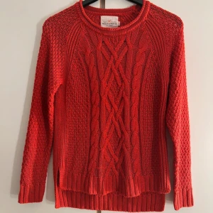 Röd stickad tröja Holly & Whyte - Riktigt snygg röd stickad tröja från Holly & Whyte med flätstickat mönster framtill och ribbade muddar. Tröjan har lång ärm, rund hals och slitsar i sidorna. Perfekt för lager på lager under hösten och vintern.