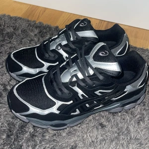 Svarta och silver Asics sneakers - Säljer ett par snygga Asics sneakers i svart och silver med mesh- och syntetpaneler. Skorna har rund tå, platt sula och coola detaljer i silver som ger dem en futuristisk vibe. Perfekta för dig som gillar sportig stil och vill sticka ut med dina skor.