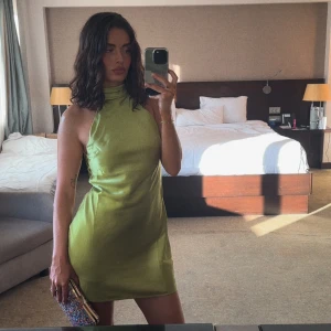 Grön axelbandslös klänning - Snygg grön axelbandslös klänning i satin med hög krage och öppen rygg. Perfekt för en kväll ute eller en speciell tillställning. Klänningen har en kort längd och en elegant passform. Använt endast en gång.