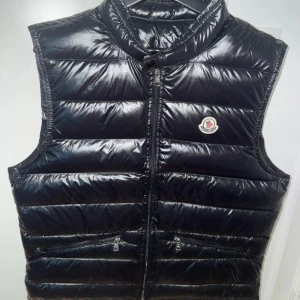 Moncler gui väst  - Snygg svart dunväst från Moncler med glansig finish och klassisk logga på bröstet. Västen har hög krage, två fickor med dragkedja och stängs med dragkedja framtill. Perfekt för lager-på-lager och streetstyle. Helt ny 