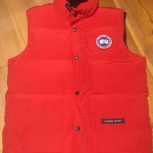 Röd dunväst från Canada Goose XS - Säljer en röd dunväst från Canada Goose i storlek XS. Västen har hög krage, tryckknappar framtill och två stora fickor. Klassisk Canada Goose-logga på bröstet och etikett på fickan. Perfekt för kalla dagar och snygg att styla över hoodie. Pris går att diskutera 