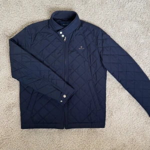 Mörkblå quiltad jacka från GANT - Säljer en stilren mörkblå quiltad jacka från GANT i storlek M. Jackan har ståkrage med tryckknappar, dragkedja framtill och två fickor. Jackan är använd en säsong men är i så gott som nyskick (se krage på bild 4). Nypris: 2500 kr