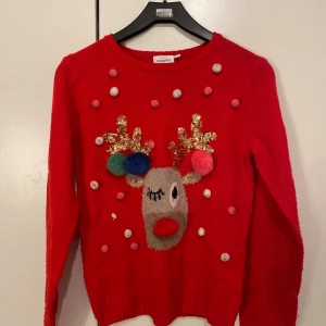 Röd jultröja med renmotiv och paljetter - Superfin röd stickad jultröja från KappAhl med ett gulligt renmotiv framtill, glittriga guldpaljetter på hornen och färgglada bollar. Tröjan har rund hals och långa ärmar, perfekt för julens alla tillfällen!