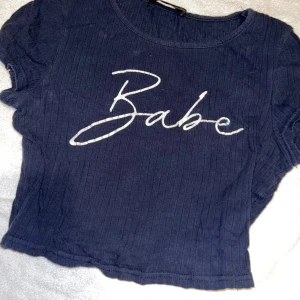 Ribbad topp - Säljer en mörkblå croppad topp med vit text 'Babe' framtill. Toppen är ribbad och har korta ärmar. Perfekt för dig som gillar en enkel men trendig stil. Finns små fläckar som jag inte provat att få bort
