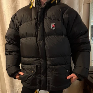 Svart dunjacka från Fjällräven XS - Säljer en svart dunjacka från Fjällräven i herrmodell, storlek XS. Nytt skick och inga defekter. Jackan har broderad logga på bröstet, dragkedja och tryckknappar framtill. Puffig och varm modell med klassisk look, perfekt för kalla dagar. 
