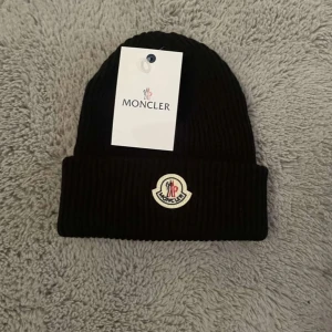 Svart ribbad mössa från Moncler - Svart ribbstickad mössa från Moncler med klassisk uppvikt kant och broderad logotyp framtill. Snygg och stilren design som passar perfekt till kalla dagar. Mössan är tillverkad i mjukt material och har en exklusiv känsla.