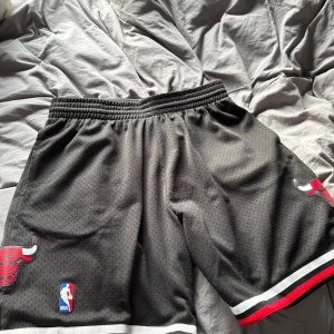 Chicago Bulls basketshorts svart - Svarta Chicago Bulls basketshorts med elastisk midja och ventilerande meshmaterial. Klassisk Bulls-logga på båda benen och NBA-märke framtill. Ränder i vitt och rött längst ner på benen ger en sportig vibe. Perfekta för basket eller chill.