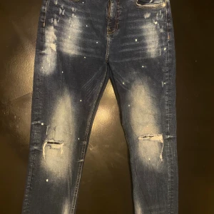 Mörkblå skinny jeans Supply & Demand - Mörkblå skinny jeans från Supply & Demand med slitningar och hål på knäna, samt vita färgstänk för en cool, streetinspirerad look. Jeansen har klassiska fem fickor och kontrastsömmar. Passar dig som gillar en trendig och avslappnad stil.