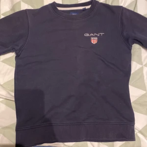 Mörkblå sweatshirt från GANT - Säljer en stilren mörkblå sweatshirt från GANT med broderad logga och sköld på bröstet. Tröjan har rund halsringning, långa ärmar och ribbade muddar vid ärmslut och nederkant. Perfekt för en clean och klassisk look.