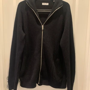 Svart stikad kofta från Jack & Jones - Svart stickad kofta från Jack & Jones med hel dragkedja framtill och hög krage. Tröjan har långa ärmar och är tillverkad i ett mjukt stickat material som passar perfekt till kyligare dagar. Enkel och stilren design som funkar till det mesta.