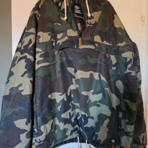 Camouflage vindjacka från Dickies XL - Säljer en oversized vindjacka från Dickies i storlek XL med klassiskt camouflage-mönster. Jackan har huva med snörning, stor ficka framtill och half zip-dragkedja. Perfekt för dig som gillar streetwear och vill sticka ut.
