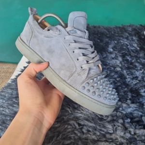 Gråa mockasneakers med nitar Louboutin - Säljer ett par gråa sneakers i mocka från Christian Louboutin med ikonisk röd sula och coola nitar framtill. Skorna har snörning och en platt sula, perfekt för dig som vill sticka ut med din stil.