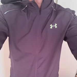 Grå vindjacka från Under Armour - Säljer en grå vindjacka från Under Armour med huva och dragkedja hela vägen. Jackan har reflekterande detaljer och logga på bröstet samt Under Armour-tryck nedtill. Perfekt för träning utomhus och har en sportig, clean look.