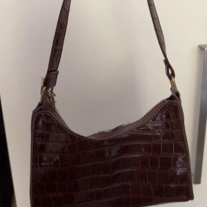 Brun axelväska i krokopräglat skinn - Snygg brun axelväska med krokopräglat mönster och glansig. Väskan har en bred axelrem och stängs med dragkedja upptill. 