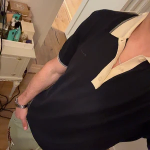 Mörkblå pikétröja från GANT - Snygg mörkblå pikétröja från GANT med kontrasterande ljusgul krage och knappslå. Klassisk passform med korta ärmar och diskret GANT-logga på bröstet. Perfekt för en stilren och avslappnad look. Aldrig använd endast testad för bilden.