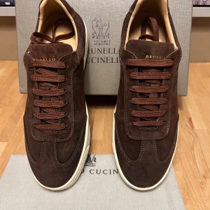 Brunello Cucinelli bruna mockasneakers - Snygga sneakers från Brunello Cucinelli i mörkbrun mocka med matchande bruna snören och vit platt sula. Skorna har rund tå och diskret guldfärgad logga på hälen. Perfekta för dig som gillar stilrena och exklusiva sneakers med lyxig känsla.