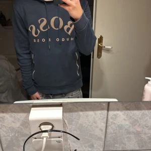 Mörkblå hoodie från Hugo Boss - Säljer en snygg mörkblå hoodie från Hugo Boss med stor logga i beige framtill. Tröjan har huva med snörning, ribbade muddar och dragkedjor vid fickorna. Insidan av huvan är beige för en schysst kontrast. Perfekt för dig som gillar stilrena streetwear-plagg. Passar 170 och under