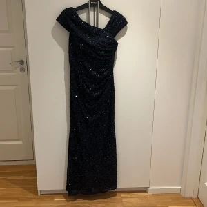 Mörkblå glittrig långklänning - Säljer en elegant mörkblå långklänning från McGregor med glittriga paljetter över hela klänningen. Den har offshoulder-skärning och figurnära passform som ger en snygg siluett. Perfekt för bal eller festliga tillfällen när du vill sticka ut.