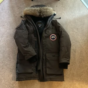 Brun Canada Goose parkas med päls - Säljer en brun Canada Goose parkas med stor huva och äkta pälsdetalj. Jackan har flera praktiska fickor, klassisk logga på bröstet och tydlig Canada Goose-märkning på ryggen. Perfekt för kalla vinterdagar och riktigt snygg streetstil.