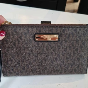 Brun plånbok från Michael Kors - Snygg brun plånbok från Michael Kors med klassiskt MK-mönster och guldfärgad logoplatta. Plånboken har flera kortfack, fönster för ID och dragkedja. Utsidan är i mönstrad canvas och insidan i svart skinn. Perfekt för att hålla koll på alla kort och cash.