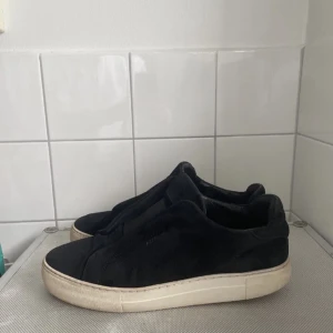 Svarta sneakers från Axel Arigato - Stilrena svarta sneakers från Axel Arigato i mocka med vit platt sula. Skorna har minimalistisk design utan snörning och elastisk panel över vristen för enkel påtagning. Perfekta för dig som gillar en clean och modern look.
