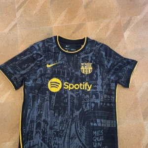 Svart fotbollströja - Snygg fotbollströja i svart med detaljer i guld och tryck. Tröjan har korta ärmar, klubbmärke och sponsorloggor på bröstet samt ett unikt stadsmotiv över hela tyget. Perfekt för dig som vill sticka ut på planen eller läktaren.