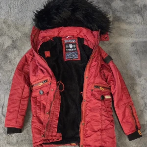 Röd parkasjacka från Navahoo S - Snygg röd parkasjacka från Navahoo med svart fuskpäls på huvan och mysigt svart fleecefoder. Jackan har flera fickor med dragkedjor och knappar, samt ribbade muddar vid ärmsluten. Perfekt för kalla dagar och har en riktigt urban vibe.