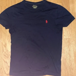 Svart t-shirt Polo Ralph Lauren XS - Snygg svart t-shirt från Polo Ralph Lauren i bomull. Klassisk röd broderad logga på bröstet och slim fit passform. Perfekt basic-plagg med korta ärmar och rund hals. Passar dig som gillar stilren och tidlös design.
