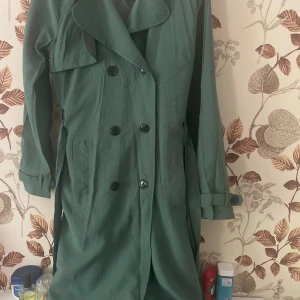 Grön trenchcoat med knappar - Snygg grön trenchcoat med dubbelknäppning och bred krage. Jackan har långa ärmar med spänne vid ärmslut och två stora fickor framtill. Perfekt för dig som vill ha en stilren och trendig look.