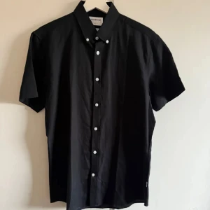 Skjorta - Snygg svart kortärmad skjorta från Lindbergh i slim fit. Tillverkad i en skön blandning av bomull och linne, perfekt för en stilren look. Skjortan har vita knappar som ger en fin kontrast mot det svarta tyget.