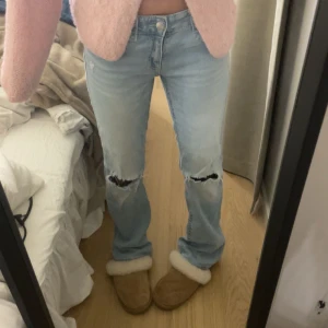 Ljusblå jeans med slitna knän - Säljer ett par ljusblå jeans med raka ben och slitna detaljer vid knäna. Jeansen har klassisk femficksdesign och snygga bakfickor med lock och knapp. De är low waist och har en avslappnad passform. Perfekta för en chill och trendig look.