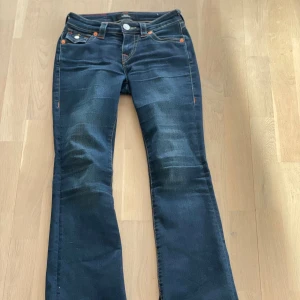 True Religion Becca Bootcut Jeans - Mörkblå bootcut jeans från True Religion, modell Becca Mid Rise. Klassiska femficksjeans med kontrastsömmar, snygga detaljer på bakfickorna och silverfärgade knappar. Jeansen är tillverkade i stretchig denim för en bekväm passform.