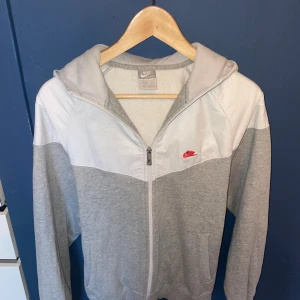 Nike grå och vit vindjacka med huva - Nike vindjacka i grått och vitt med huva och hel dragkedja framtill. Jackan har en snygg tvåfärgad design, röd Nike-logga på bröstet. Materialmix med mjuk bomull och syntet. Den ska va S men passformen e lite loose så hade kunnat funka dom M