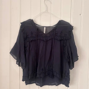 Mörkblå blus med volang från Pepe Jeans - Supersöt mörkblå blus från Pepe Jeans med broderade detaljer och volanger längs ärmar och bröst. Blusen är luftig med v-ringning, knytband med tofsar och vida trekvartsärmar. 