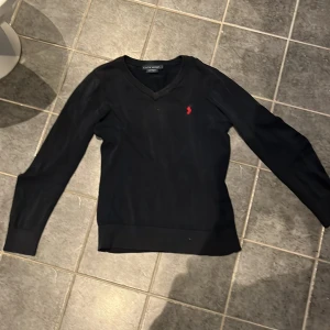 Marinblå  v-ringad tröja Ralph Lauren L - Marinblå v-ringad tröja från Ralph Lauren med röd broderad logga på bröstet. Tröjan är långärmad och har ribbade muddar vid ärmslut och nederkant. Custom fit-modell som sitter snyggt och passar perfekt till lager-på-lager-stil. OBS  BARNSTORLEK!!