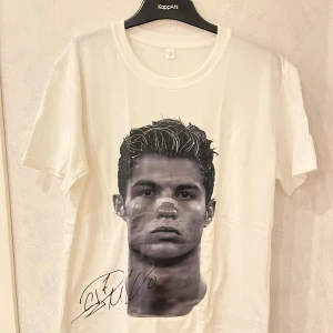 Ronaldo T-shirt  - Vit t-shirt från KappAhl i bomull med coolt svartvitt tryck av ett huvud framtill och en tryckt autograf nedtill. Klassisk rund halsringning och korta ärmar. Perfekt för dig som gillar grafiska motiv och enkel stil.
