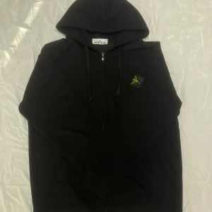 Stone Island Hoodie Zip - Svart hoodie från Stone Island med dragkedja framtill och klassisk logga på vänster ärm. Tröjan har huva med snörning och ribbade muddar. Perfekt för dig som gillar streetwear och vill ha en stilren look.