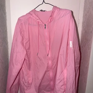 Rosa vindjacka  - Säljer en rosa vindjacka från RISKY med huva och vit logga på ärmen samt stort tryck på huvan. Jackan har dragkedja framtill, snörning i huvan och är tillverkad i lätt polyester. Perfekt för dig som vill sticka ut med färg och streetstyle.