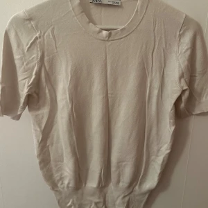 Vitt stickad kortärmad tröja Zara - En stilren beige stickad tröja från Zara med rund halsringning och korta ärmar. Tröjan har en enkel och clean look som passar perfekt till jeans eller kjol. Mjuk och skön kvalitet som känns lyxig mot huden.