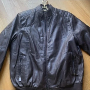 Brun skinnjacka med dragkedja - Snygg brun skinnjacka i klassisk bombermodell med ribbade muddar och ståkrage. Jackan har dragkedja framtill och två fickor med dragkedja. Perfekt för dig som vill ha en stilren och tidlös look.