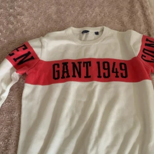 Vit och röd stickad tröja GANT - Vit stickad tröja från GANT med röd bred rand över bröstet och armarna, samt svart text 'GANT 1949'. Rund halsringning och långa ärmar. Perfekt för dig som gillar klassisk och sportig stil.