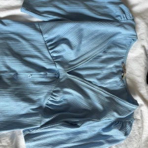 Ljusblå topp  från Stradivarius - Supersöt ljusblå klänning från Stradivarius i storlek S. Klänningen har v-ringning, knappar framtill och puffiga korta ärmar. Materialet är ribbat och ger en snygg struktur. Perfekt för dig som gillar en feminin och trendig look.