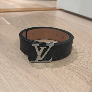 Bälte - Snyggt svart bälte med ikoniskt spänne i silverfärgad metall. Bältet har diskret monogrammönster och är tillverkat i exklusivt skinn med ljusbrun insida. Perfekt accessoar för att lyfta din outfit.