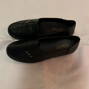 Svarta loafers med pärldetaljer - Stilrena svarta loafers med rund tå och diskreta pärldetaljer framtill. Skorna har en klassisk design och är tillverkade i syntetmaterial med en platt sula för bekväm användning hela dagen.